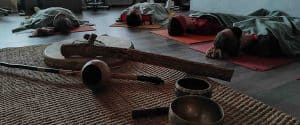 Inquadratura ravvicinata di campane tibetane, tamburo sciamanico e rainstick disposti su una stuoia chiara con partecipanti distesi a terra coperti da coperte durante un bagno sonoro di purificazione al Centro Zorba Il Buddha.
