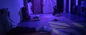 Partecipanti seduti e distanziati a terra in una sala illuminata da luce viola soffusa con le mani sul capo o sul volto durante la meditazione porta la luce nei chakra, tecniche di meditazione Bodhi Vipal al Centro Zorba Il Buddha.