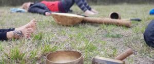 Partecipanti distesi a piedi nudi sull'erba durante un ritiro di Shinrin-Yoku con campane tibetane e strumenti sonori sciamanici all'aperto, tecniche di meditazione Bodhi Vipal al Centro Zorba Il Buddha.