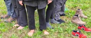 Primo piano dei piedi di più persone in fila su un prato verde, con diverse opzioni per il contatto con il suolo: a piedi nudi, calzini e scarpe rimosse. Pratica di meditazione Bodi Vipal, Centro Zorba Il Buddha