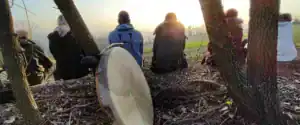 Tamburo sacro sciamanico appoggiato a un albero in primo piano con praticanti seduti di spalle a contemplare il tramonto al Centro Zorba Il Buddha.