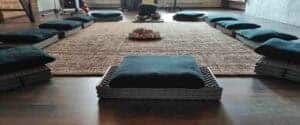 Cuscini zafu scuri e materassini disposti in cerchio attorno a un grande tappeto con campane tibetane nella sala da meditazione Buddha Hall del Centro Zorba Il Buddha.