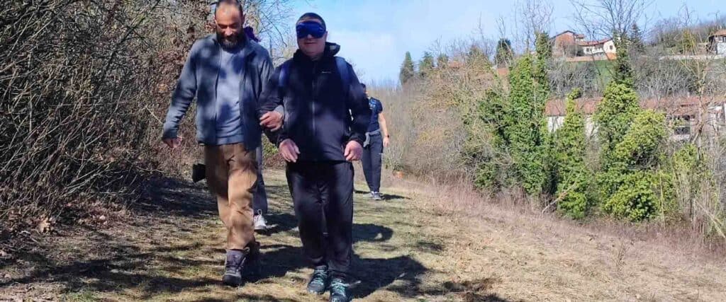 : Un uomo con una benda sugli occhi cammina lungo un sentiero collinare, guidato a braccetto da un compagno durante un'attività di escursione sensoriale.