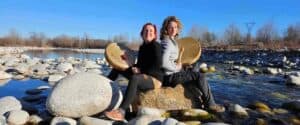 Due donne sedute schiena contro schiena su una roccia in mezzo a un fiume cristallino, mentre suonano tamburi sciamanici durante un seminario di Costellazioni Familiari.