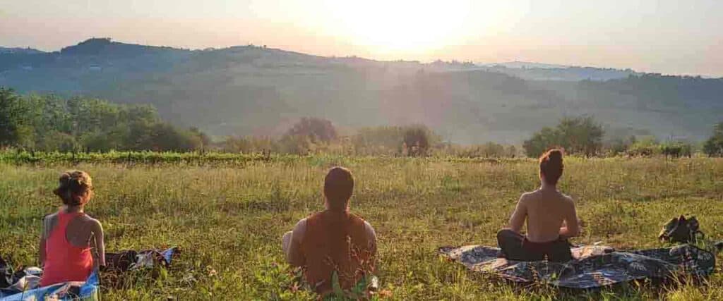 Tre persone sedute in meditazione su un prato all'alba di fronte a un paesaggio collinare.