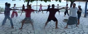 Un gruppo di persone esegue in cerchio la posizione yoga del Guerriero II (Virabhadrasana II) tenendosi per mano su una terrazza panoramica in pietra con vista sul mare, durante un ritiro al Centro Zorba Il Buddha.