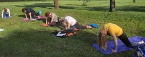 Gruppo di persone su tappetini colorati in un prato verde mentre eseguono la posizione yoga equestre (Ashwa Sanchalanasana) durante una sessione all'aperto al Centro Zorba Il Buddha.